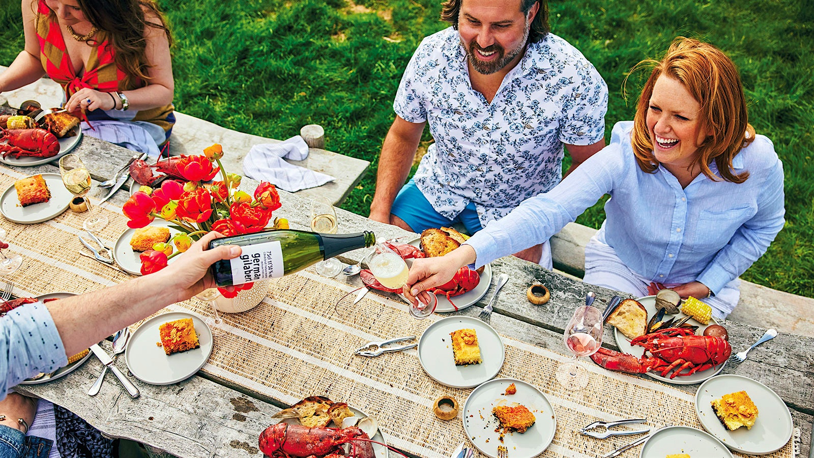 Three people enjoy a lobster boil at a table; at left, a man pours a glass of sparkling wine.